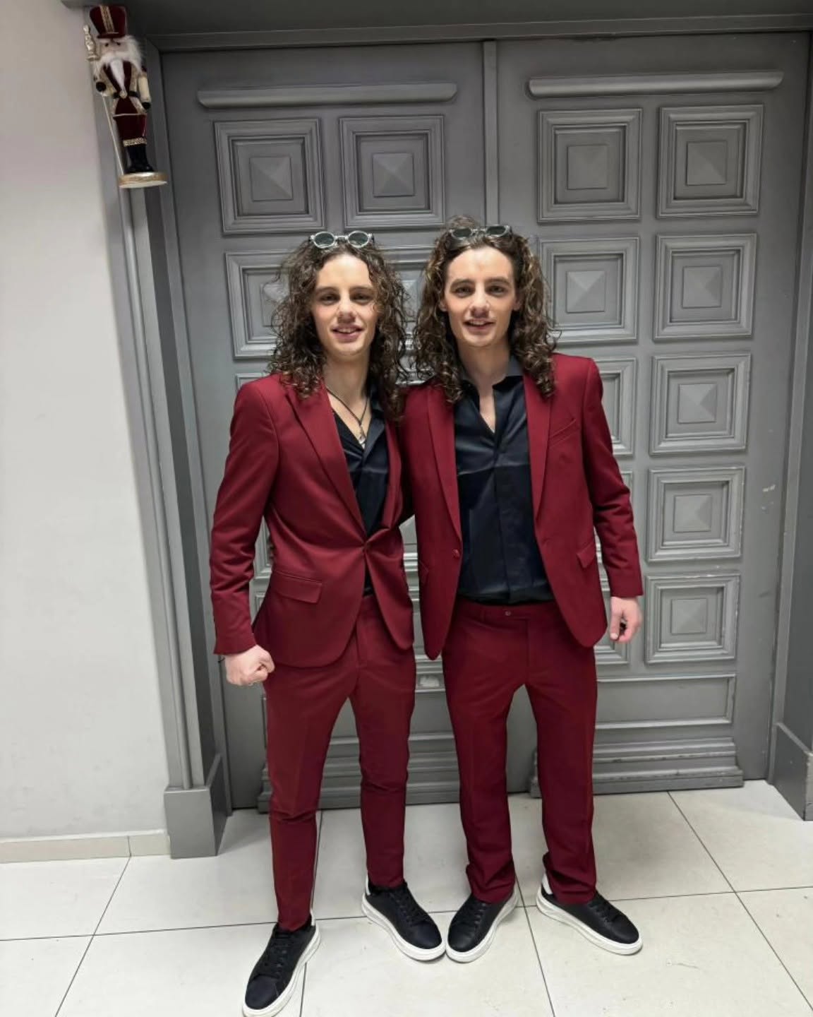 Two people stand side by side wearing matching burgundy suits and black shirts, framed full-length against a paneled gray door.