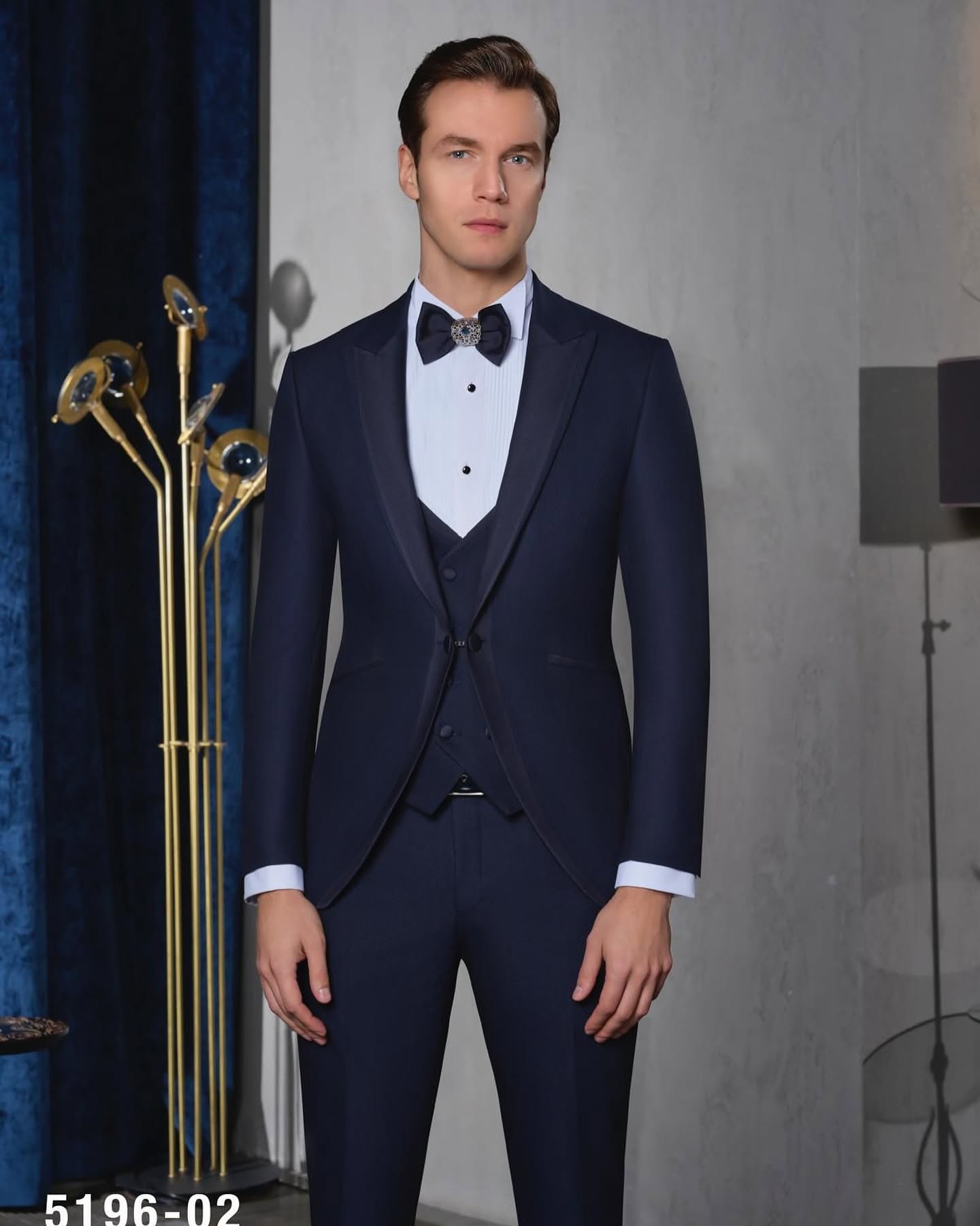 Studio-style portrait of a man wearing a navy tuxedo, standing against a dark interior backdrop with soft lighting.