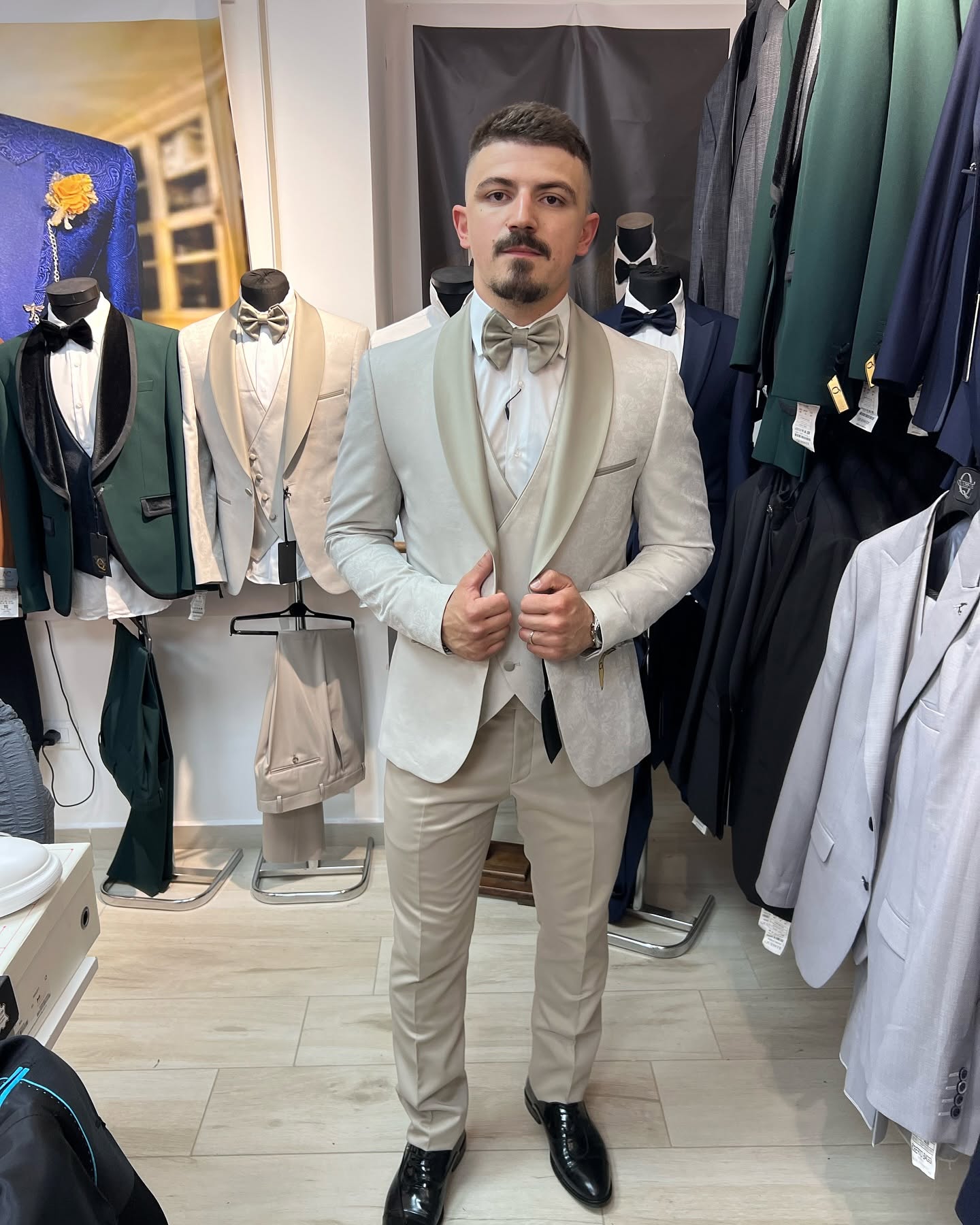 A man stands full-length wearing a light-colored tuxedo with a bow tie, surrounded by hanging jackets and mannequins in bright indoor lighting.