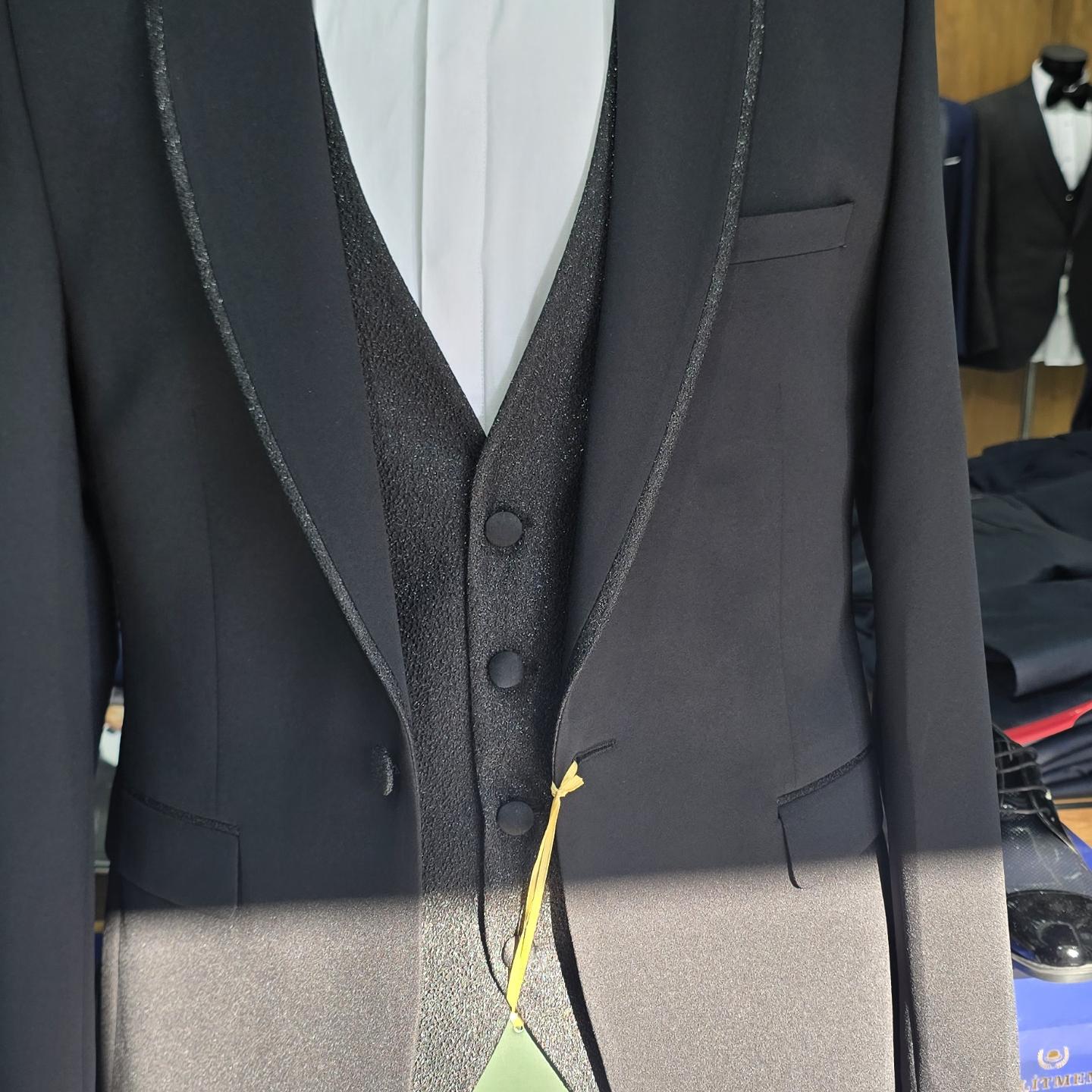 Close-up photograph of a black formal suit jacket with contrasting lapels and vest, framed from the chest to the waist under bright indoor lighting.