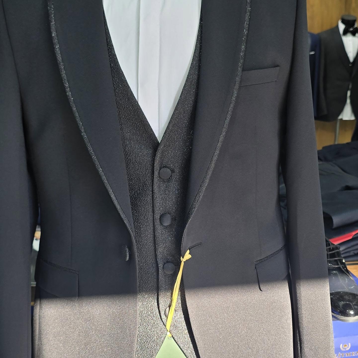 A close-up shows the front of a black tuxedo jacket, vest and white shirt with textured lapels and a hanging yellow tag.