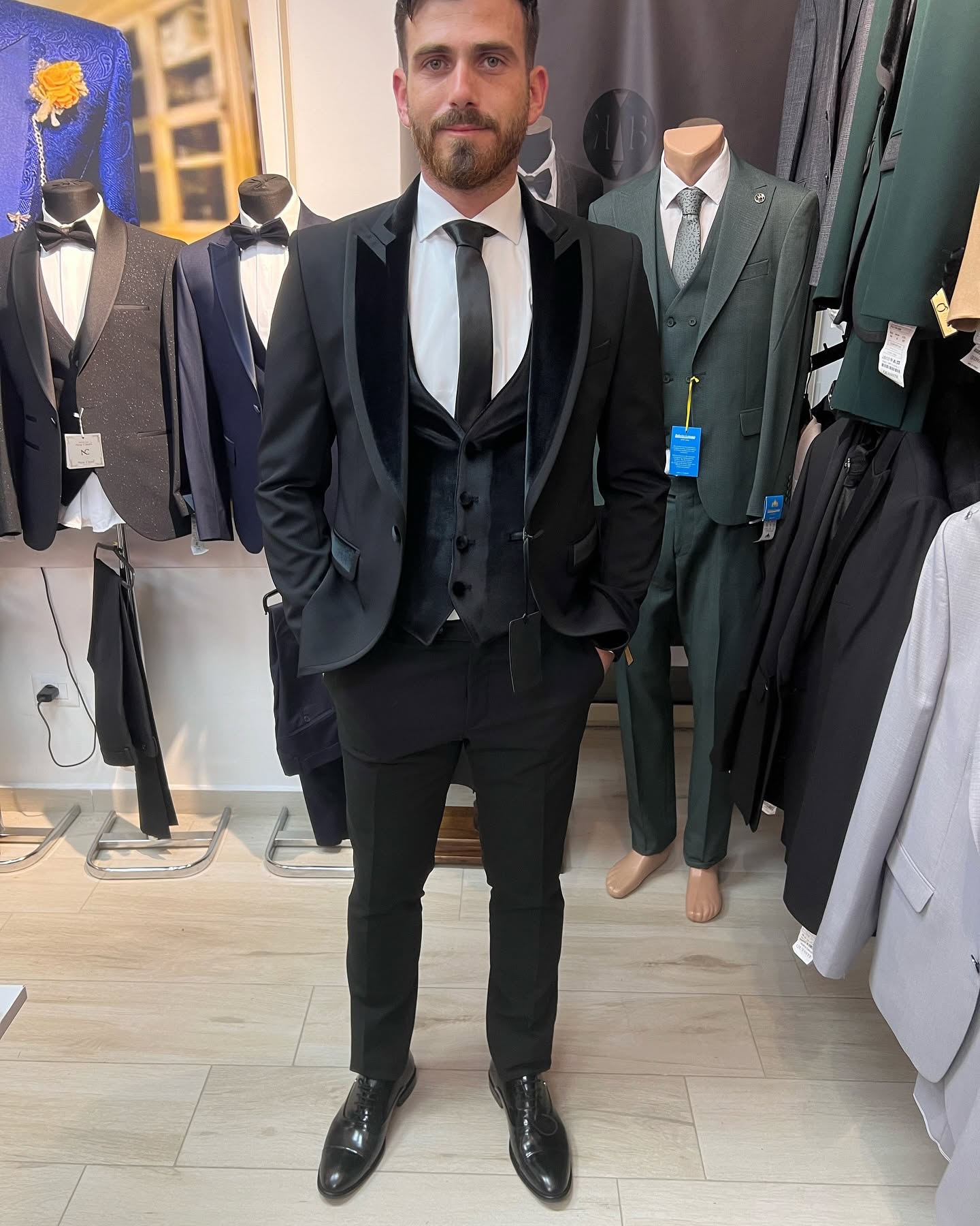 Full-body view of a man wearing a black tuxedo in a showroom, with clothing racks and mannequins in the background.