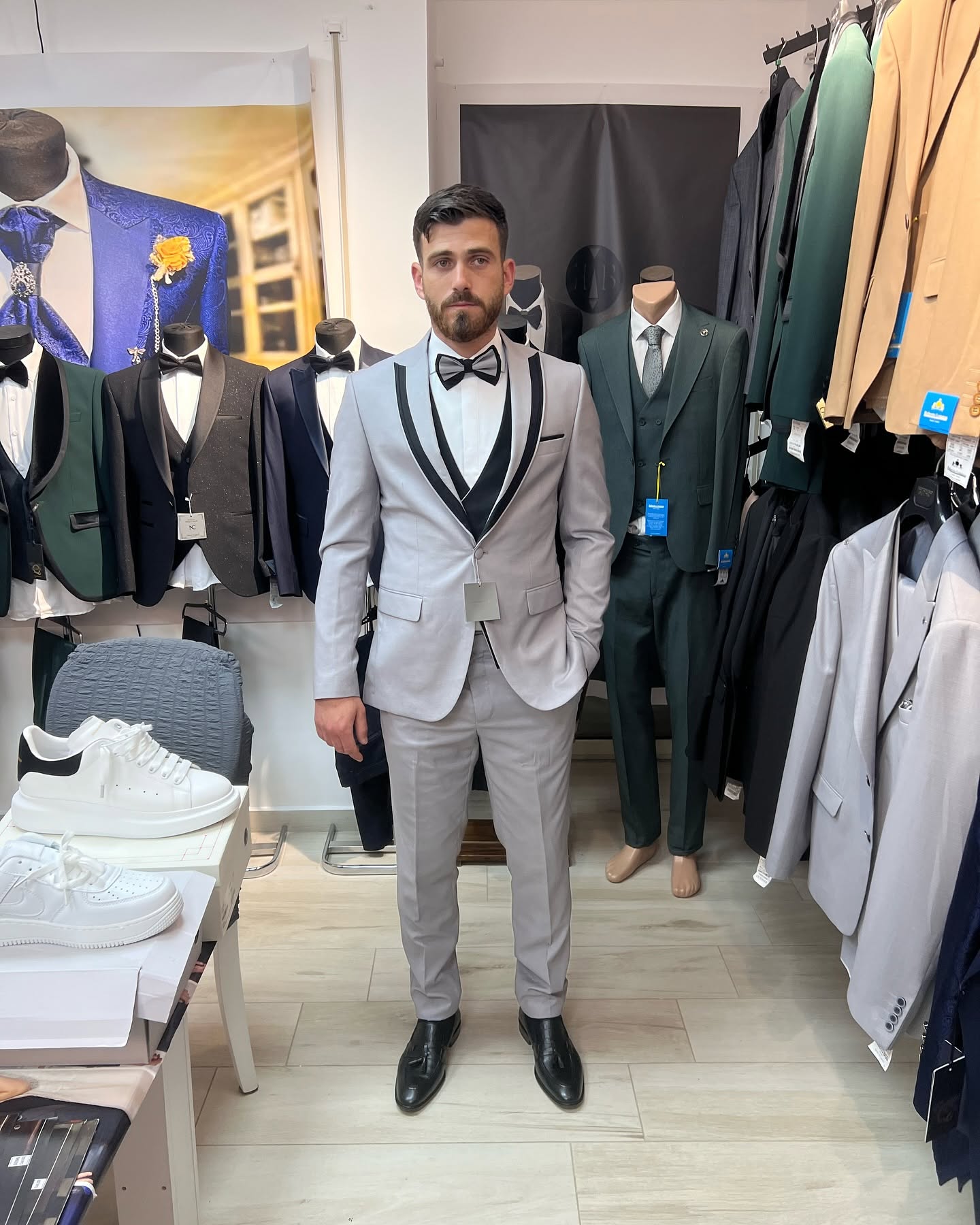 Full-body view of a man wearing a light gray tuxedo inside a clothing showroom, with suits and mannequins surrounding him under indoor lighting.