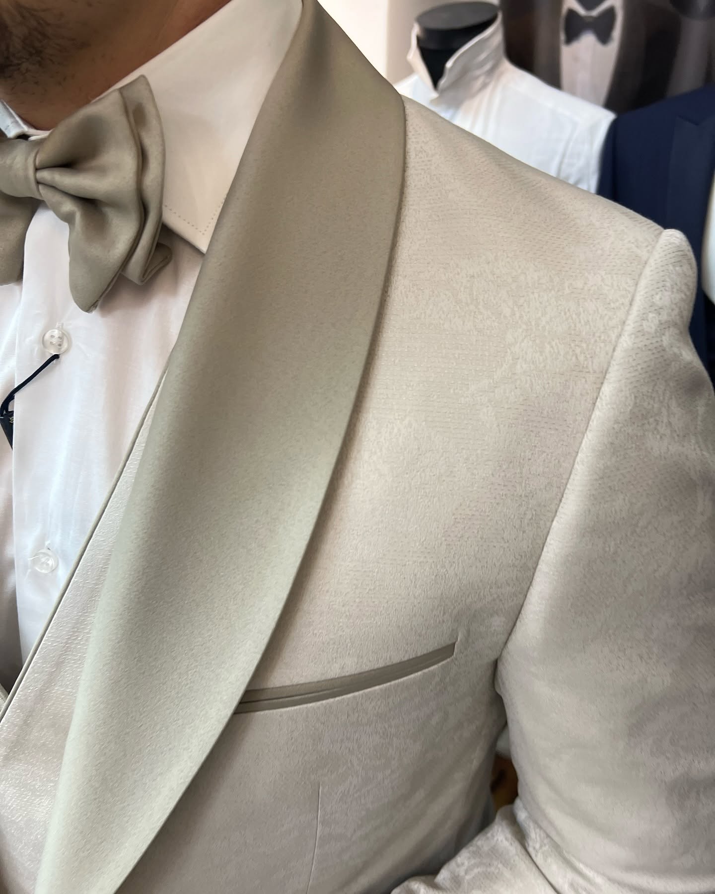 Close-up of a cream-colored tuxedo jacket and bow tie, showing the lapel and pocket detail under indoor lighting.