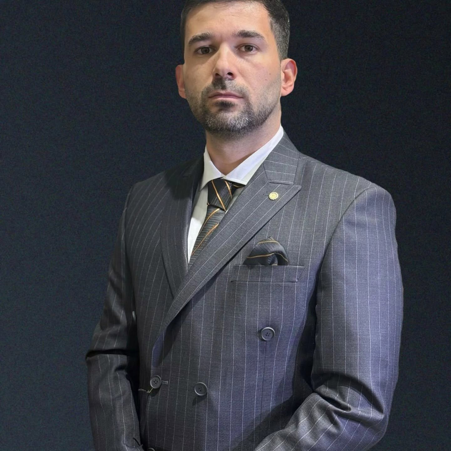 A man is shown from the chest up wearing a gray pinstriped double-breasted suit against a dark background with directional lighting.