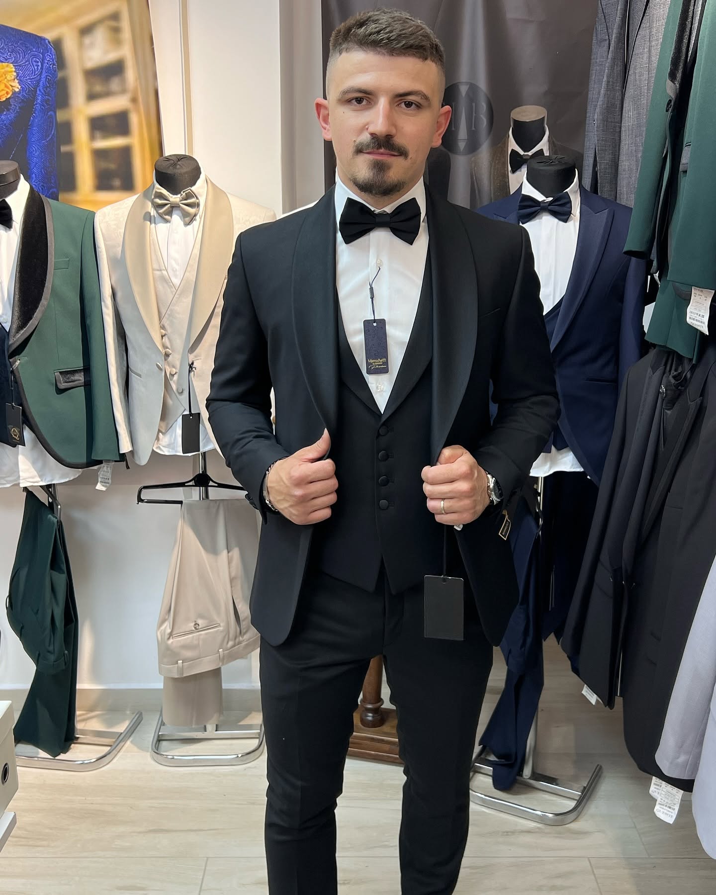 Full-body view of a man wearing a black tuxedo in a showroom, surrounded by displayed suits and racks under indoor lighting.