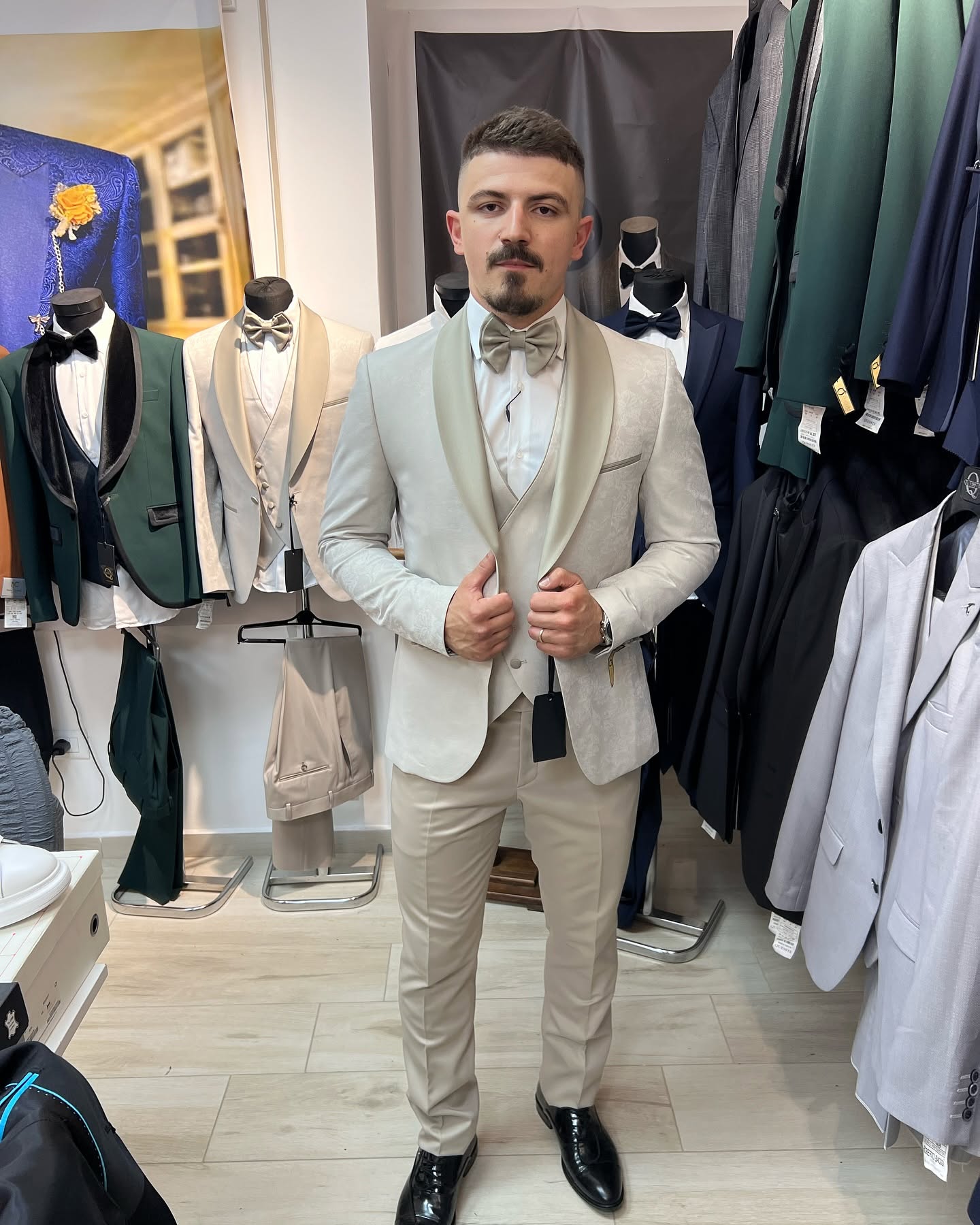 Full-body view of a man wearing a cream-colored tuxedo in a showroom, with mannequins and clothing racks behind him.