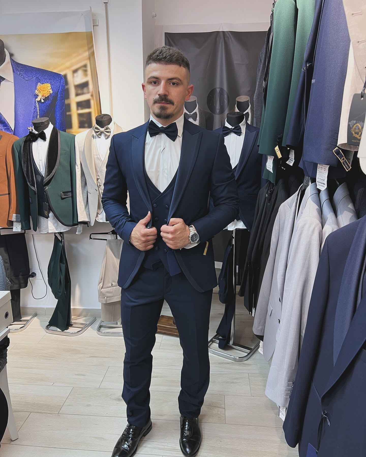 A man stands full-length wearing a navy tuxedo with a bow tie, with clothing racks and mannequins around him under bright indoor lighting.