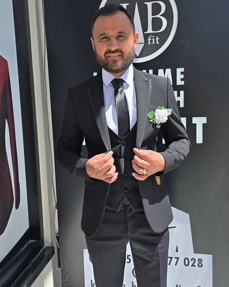 A man stands wearing a gray-and-black tuxedo with a boutonniere, photographed in bright light against a dark backdrop with visible lettering.