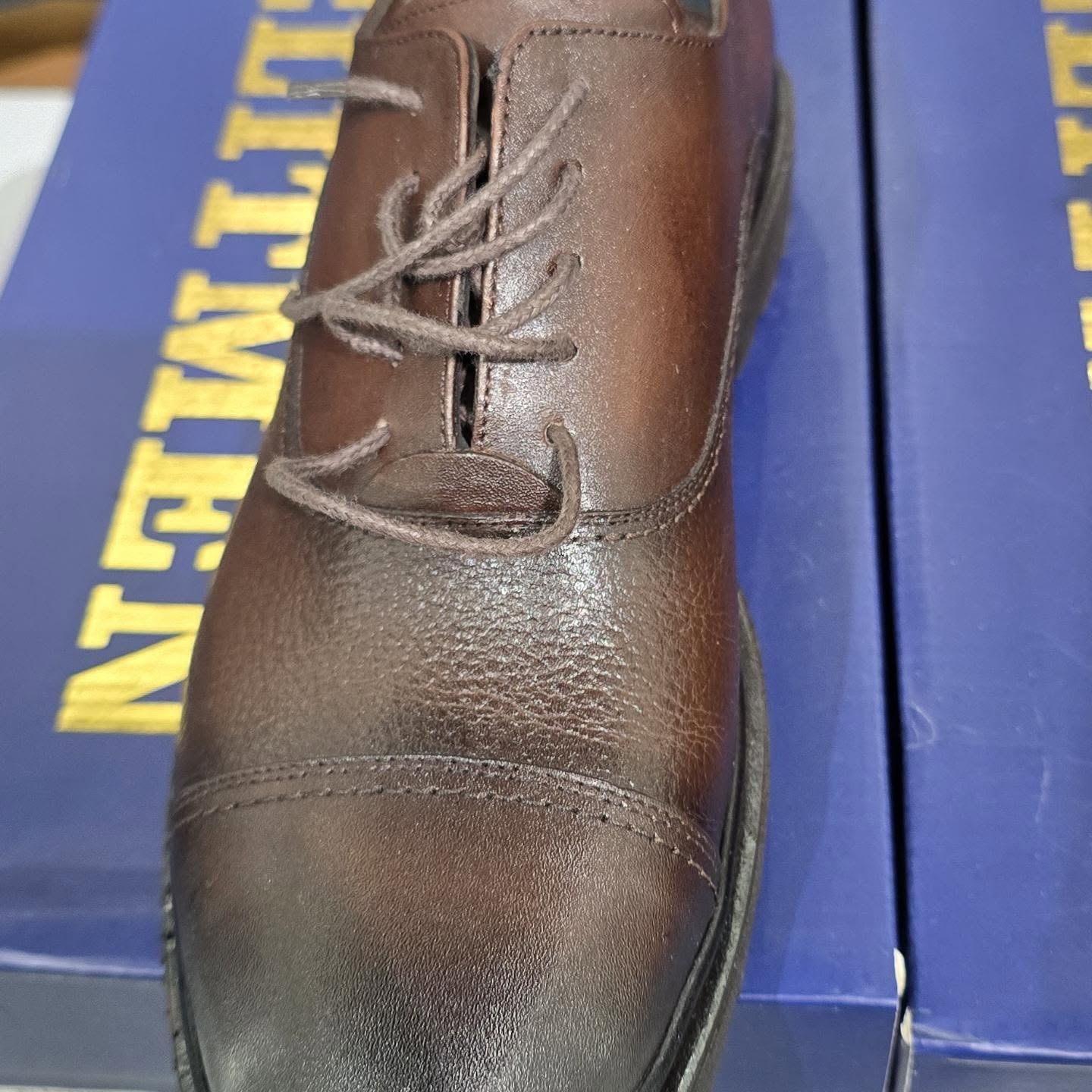 A close-up shows a brown leather lace-up dress shoe resting on blue boxes under bright lighting.